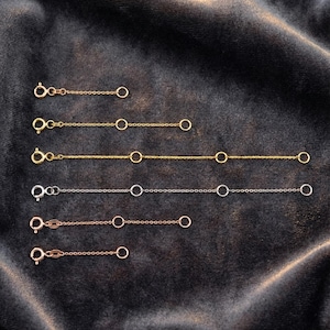 May include: A collection of gold, silver, and rose gold chain extenders with lobster claw clasps and small circular connectors. The extenders are displayed on a dark gray velvet surface.
