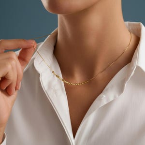Op de afbeelding: Een delicate gouden ketting met een reeks kleine, gelijkmatig verdeelde kralen. De ketting wordt gedragen tegen een wit overhemd met kraag, met een hand die de ketting zachtjes vasthoudt. De achtergrond is effen, gedempt blauw.
