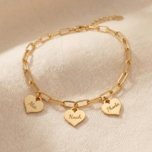 Op de afbeelding: Goudkleurige armband met een schakelkettingontwerp. De armband heeft drie hartvormige bedels, elk gegraveerd met een naam: Mia, Noah en Phoebe. De armband wordt gepresenteerd op een crèmekleurig oppervlak.