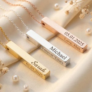Può includere: Tre collane personalizzate con barre in oro, argento e oro rosa. Ogni barra è incisa con un nome, una data e un messaggio. Le collane sono esposte su un tessuto color crema con perle e piccoli fiori bianchi.