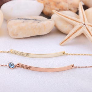 Puede incluir: Dos delicadas pulseras doradas y oro rosa. La pulsera dorada está inscrita con "NO KINGS". La pulsera de oro rosa presenta una piedra preciosa azul y la palabra "RESIST". Las pulseras se exhiben sobre una superficie blanca con conchas marinas en el fondo.