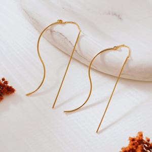 Könnte beinhalten: Ein Paar goldfarbene Ohrringe. Einer hat ein wellenförmiges, geschwungenes Design, der andere eine gerade Linie. Beide sind durch eine zarte Kette verbunden. Die Ohrringe werden auf einer weißen Oberfläche präsentiert.