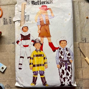 Puede incluir: Un sobre de patrón de costura Butterick vintage con ilustraciones de disfraces infantiles. Los disfraces incluyen un traje espacial, un atuendo de vaquera, un uniforme de bombero y un conjunto de vaquero. El sobre es blanco con el número 26.