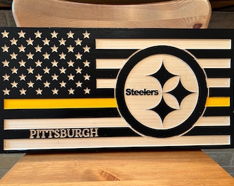 Puede incluir: Letrero de madera con el diseño de la bandera estadounidense, una franja amarilla, el logotipo de los Steelers de Pittsburgh y la palabra "PITTSBURGH". El letrero es de madera con vetas naturales y detalles en negro y amarillo.
