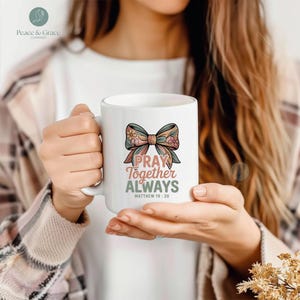Puede incluir: Taza de cerámica blanca con un diseño de lazo floral y la frase "PRAY TOGETHER ALWAYS", con Mateo 18:20. La taza es sostenida por una persona.