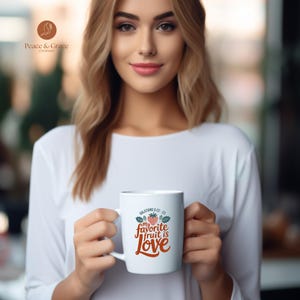 Puede incluir: Taza de cerámica blanca con el texto "my favorite fruit is Love" en una fuente cursiva, con una ilustración de fresa y hojas. La taza también tiene el texto "Galatians 5:22-23". La taza es sostenida por una persona que lleva una camisa blanca de manga larga.