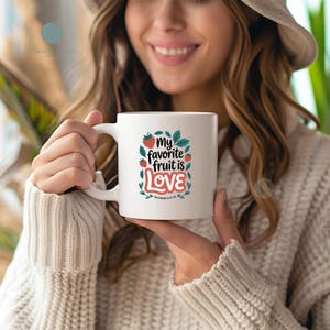 Puede incluir: Taza de cerámica blanca con el texto "My favorite fruit is LOVE" en un diseño colorido con fresas y hojas. La taza tiene un asa blanca y la sostiene una persona que lleva un suéter.