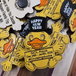 May include: A collection of yellow duck-shaped magnets. One magnet features a duck wearing a black top hat with "HAPPY NEW YEAR" on it. The duck holds a sign that reads "NCL Aqua Dec 28th - Jan4th, 2026 #Cruisingducks". Other magnets are visible.
