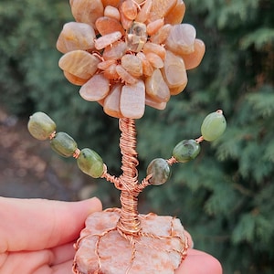 May include: A handcrafted gemstone tree sculpture. The tree features a flower-shaped top made of orange and tan stones, a copper wire trunk, and green stone leaves. The base is a rough, light orange stone.