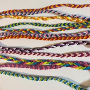 May include: A collection of colorful friendship bracelets made with braided string. The bracelets feature a variety of colors, including red, orange, yellow, green, blue, purple, pink, and white.