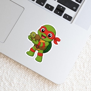 May include: A die-cut sticker featuring a cartoon green turtle character wearing a red eye mask, red wristbands, and a brown belt with a circular buckle displaying the letter R, while holding a small brown turtle in its hands.
