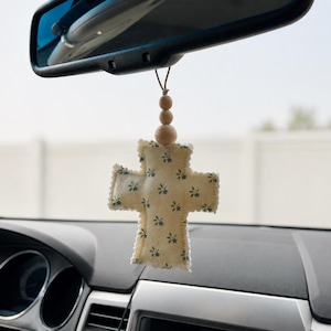 May include: A hanging car ornament featuring a cream-colored fabric cross with a small blue floral pattern. The cross has a scalloped edge and is suspended by a thin cord adorned with three round, natural-toned wooden beads hanging from a mirror.