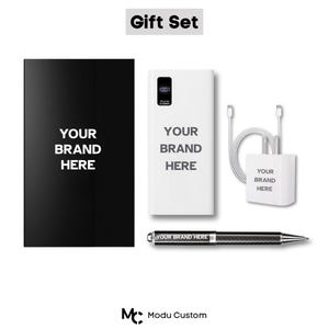 Peut inclure: Un coffret cadeau comprenant un bloc-notes noir, une batterie externe blanche avec un écran numérique, un chargeur blanc et un stylo. Chaque article affiche le texte "YOUR BRAND HERE". L'ensemble est étiqueté "Gift Set".
