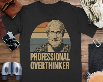 Peut inclure: T-shirt noir avec une illustration de style vintage d'un buste d'homme barbu, sur fond de coucher de soleil rétro. Le texte "PROFESSIONAL OVERTHINKER" est imprimé en dessous. Le t-shirt est présenté sur une surface en bois.