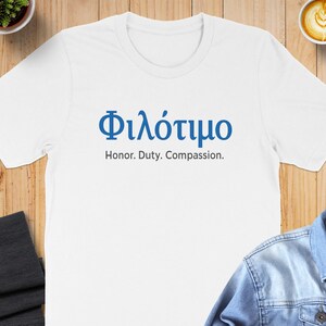Puede incluir: Camiseta blanca con la palabra griega "Φιλότιμο" en azul, junto con la traducción al inglés "Honor. Duty. Compassion." impresa debajo. La camiseta se muestra sobre una superficie de madera.