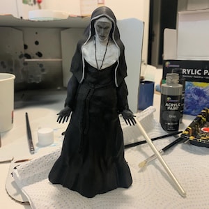 Puede incluir: Una figura de monja en blanco y negro con un cuello blanco y una túnica negra. La figura está de pie sobre una superficie blanca con pinceles y botellas de pintura cerca. Las botellas de pintura están etiquetadas como "Pintura acrílica" y "Baya madura".