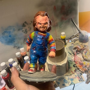 Può includere: Una statuetta dipinta a mano di un personaggio horror immaginario, un bambino con una camicia a righe rosse e blu, una salopette blu e un'espressione minacciosa. La statuetta è in piedi su una base rotonda.