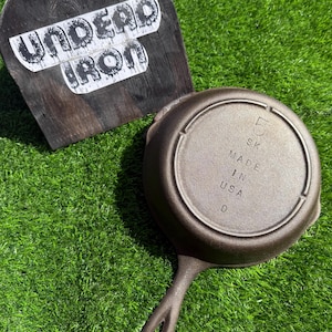May include: A cast iron skillet with a handle, resting on green grass. The bottom of the skillet is visible, with the text "5 SK MADE IN USA D". A wooden sign with the text "UNDEAD IRON" is in the background.