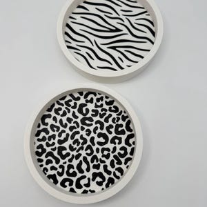 Op de afbeelding: Twee ronde witte trays met dierenprints. Eén tray heeft een zwart-wit zebrapatroon, de andere een zwart-wit luipaardprint. De trays zijn waarschijnlijk voor decoratieve doeleinden.