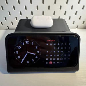 Può includere: Un supporto di ricarica nero con un pad di ricarica wireless bianco in cima. Il supporto ha un display digitale di orologio e calendario che mostra la data 28 gennaio.