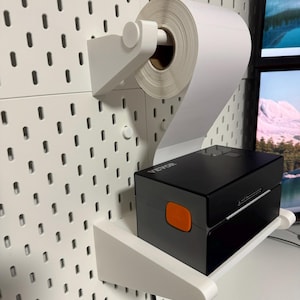 Könnte beinhalten: Eine weiße Lochwand mit einem montierten Etikettenrollenhalter und einem schwarzen Etikettendrucker. Die Etikettenrolle ist weiß und der Drucker hat eine orangefarbene Taste. Die Lochwand ist an einer Wand befestigt.