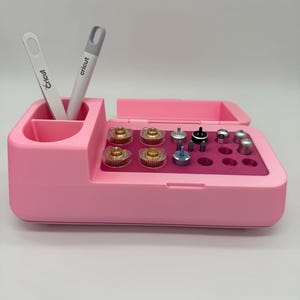 Peut inclure: Un organisateur d'outils Cricut rose avec un compartiment pour les outils et accessoires. L'organisateur a une section pour les stylos et une section pour divers embouts Cricut. Deux outils Cricut blancs et gris sont dans le porte-stylo.