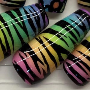 Op de afbeelding: Close-up van verschillende nail art ontwerpen. De focus ligt op drie lange, ovaalvormige nagels met een regenboog zebra streep patroon. De kleuren gaan over van roze naar geel naar groen naar blauw. Andere nagels met luipaardprint zijn zichtbaar.