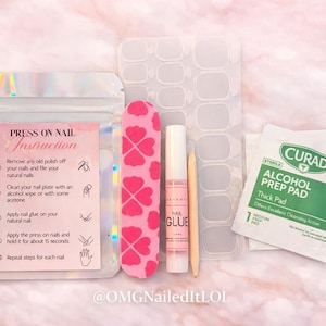 Op de afbeelding: Een pers-on-nagelkit met instructies, inclusief een roze nagelvijl met klaverpatroon, nagellijm, een bokkenpoot, nagelvoorbereidingspads en nagellijmstrips. De kit staat op een roze en witte achtergrond.