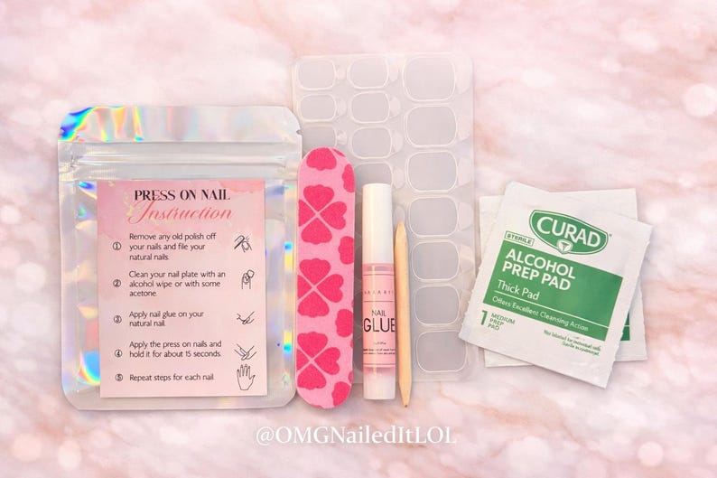 Op de afbeelding: Een pers-on-nagelkit met instructies, inclusief een roze nagelvijl met klaverpatroon, nagellijm, een bokkenpoot, nagelvoorbereidingspads en nagellijmstrips. De kit staat op een roze en witte achtergrond.
