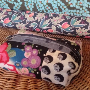 Peut inclure: Une petite pochette zippée avec un motif patchwork à motifs floraux et à pois. La pochette est posée sur une surface en osier tressé, avec des rouleaux de tissu à motifs en arrière-plan.