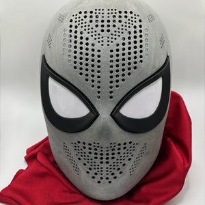 Könnte beinhalten: Eine graue Superheldenmaske mit schwarzen Augenrändern und weißen Netzlinsen. Die Maske hat zahlreiche kleine Löcher zur Belüftung und ist vor einem roten Stoffhintergrund platziert. Die Maske ist ein Cosplay- oder Kostümartikel.