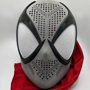 Op de afbeelding: Een grijs en zwart superheldenmasker met witte mesh oogbedekkingen en een rode stof eronder gedrapeerd. Het masker heeft een geperforeerd ontwerp op het voorhoofd en de wangen, en zwarte randen.