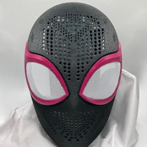Könnte beinhalten: Eine schwarze Vollmaske mit texturierter, perforierter Oberfläche und großen, weißen Netz-Augenlinsen, die in leuchtendem Pink eingefasst sind. Die Maske hat eine abgerundete Oberseite und ein spitz zulaufendes Kinn mit 3D-Druck-Struktur.