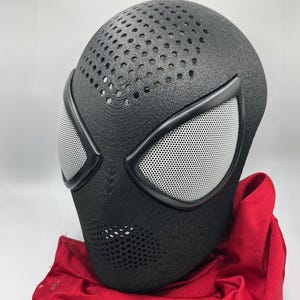 May include: A black full-face mask with a textured surface and a pattern of small circular perforations across the forehead and mouth area. The mask features two large, almond-shaped eye openings covered with a fine white mesh and a red fabric.