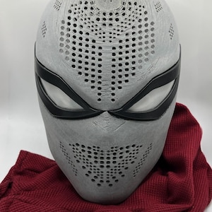 May include: A gray, full-face mask with a series of small, evenly spaced holes. The mask has black eye coverings and is resting on a red fabric. The mask appears to be 3D-printed.