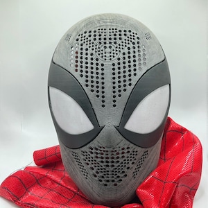 May include: A gray and black Spider-Man mask with white eye lenses and a red fabric cowl. The mask has a textured surface with numerous small holes for ventilation. The red cowl has a black web pattern.