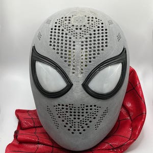 May include: A grey Spider-Man mask with black eye frames and white eye lenses. The mask has many small holes for ventilation and is paired with a red fabric cape with a black web pattern.