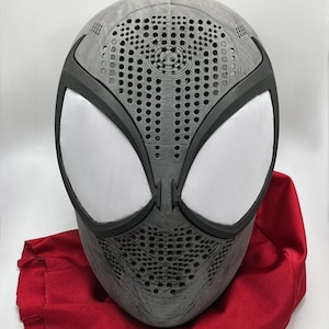 May include: A gray and black Spider-Man mask with white mesh eye coverings. The mask has a textured surface and is resting on a red cloth bag. The mask has many small holes for ventilation.