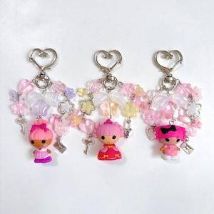 May include: Three keychains with doll figures, each featuring a heart-shaped clasp. The keychains have pink, white, and clear beads, bows, and flower accents. Each doll has a different pink hairstyle and outfit.