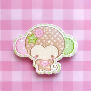 May include: A die-cut sticker featuring a stylized brown monkey character with a polka-dot head, a pink button on its forehead, a green star-shaped accent, and a small pink heart on its chest. The design includes a curly tail and glittery effects.