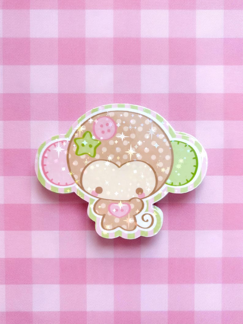 May include: A die-cut sticker featuring a stylized brown monkey character with a polka-dot head, a pink button on its forehead, a green star-shaped accent, and a small pink heart on its chest. The design includes a curly tail and glittery effects.