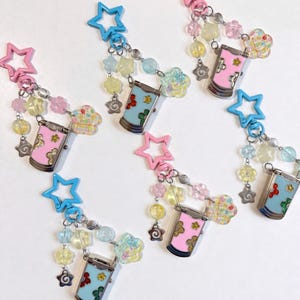 May include: Five keychains featuring a miniature flip phone charm in pink or blue with floral accents. Each keychain includes a star-shaped clasp, silver-toned metal chains, and various translucent beads in shapes like stars and flowers in pastel colors.