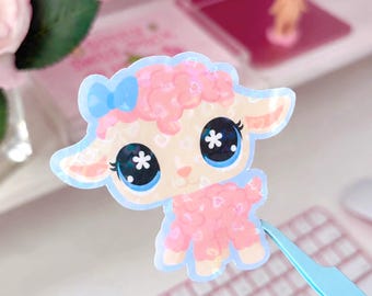 May include: A die-cut sticker featuring a cartoon lamb with pink curly wool, large blue eyes with white star highlights, and a small blue bow on its head. The design includes a subtle heart pattern overlay and a light blue border outline.