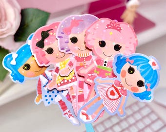 May include: A collection of five die-cut stickers featuring stylized dolls with large black button eyes, vibrant hair colors including blue and pink, and detailed outfits. The stickers have a holographic finish that reflects light with small star patterns.