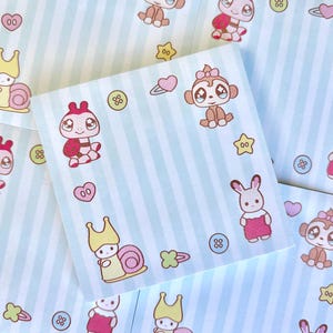 May include: A square memo pad sheet featuring a light blue and white vertical striped pattern. The design includes repeating illustrations of a brown monkey, a pink snail, a white rabbit, small buttons, hearts, and hair clips.