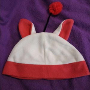 Puede incluir: Sombrero de lana blanca con ribete rojo y orejas de conejo. El sombrero tiene un pompón rojo en la parte superior.