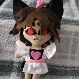 May include: A plush doll with brown hair, large black ears, and red eyes. The doll wears a white apron with ruffled sleeves over a black bodice featuring a pink heart. A pink ribbon is tied around the waist, and a pink loop is attached to the top.
