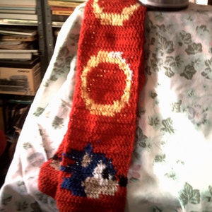 May include: A red crocheted scarf with yellow and white rings and a blue and white image of Sonic the Hedgehog.
