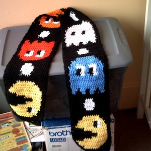 May include: A black crocheted scarf with colorful Pac-Man characters. The scarf features Pac-Man, Blinky, Pinky, Inky, and Clyde.