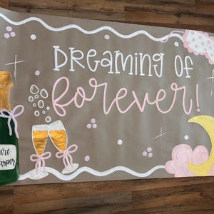 May include: A hand-painted sign with the words "Dreaming of forever!" in white and pink script. The sign features a green champagne bottle, two champagne flutes, a crescent moon, and a sleep mask. The words "Future Mrs. Harper" are on the champagne bottle.